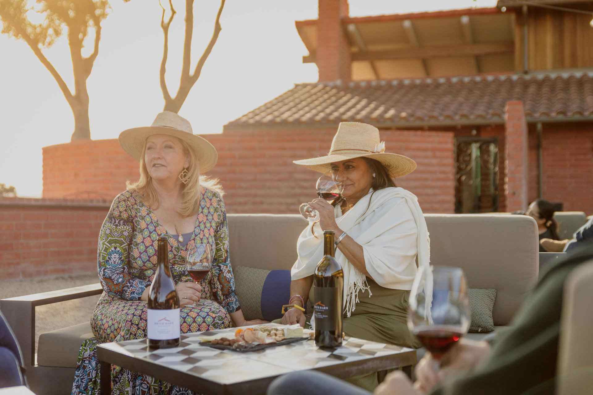 Two women with large hats sipping wine on a couch outside.
